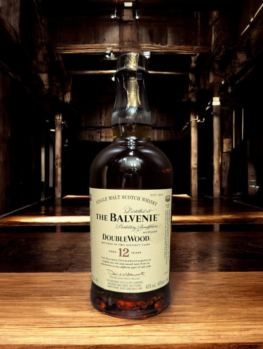 Ein stilvoll arrangiertes Foto einer Flasche Balvenie 12 Years Old DoubleWood Single Malt Scotch Whisky vor einem dunkelgrauen Hintergrund. Die honigfarbene Flüssigkeit schimmert durch das klare Glas, das mit einem cremefarbenen Etikett verziert ist. Der Name "Balvenie" und die Whisky-Details sind in eleganter Schrift abgedruckt. Die Szene vermittelt ein Gefühl von Raffinesse und Handwerkskunst, das den Charakter dieses Whiskys widerspiegelt.
