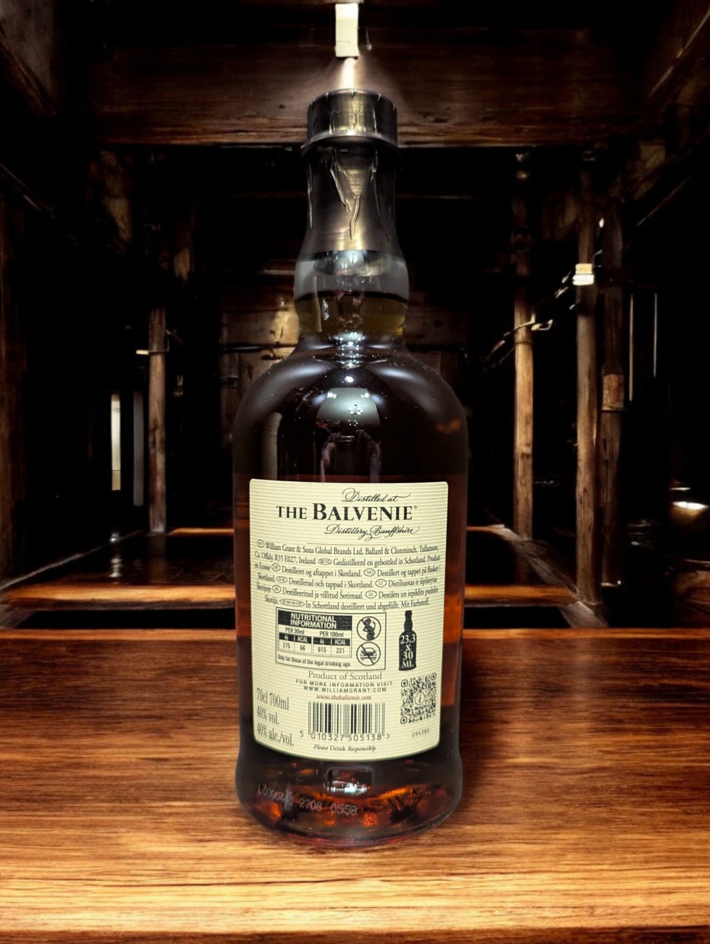 Ein stilvoll arrangiertes Foto einer Flasche Balvenie 12 Years Old DoubleWood Single Malt Scotch Whisky vor einem dunkelgrauen Hintergrund. Die honigfarbene Flüssigkeit schimmert durch das klare Glas, das mit einem cremefarbenen Etikett verziert ist. Der Name "Balvenie" und die Whisky-Details sind in eleganter Schrift abgedruckt. Die Szene vermittelt ein Gefühl von Raffinesse und Handwerkskunst, das den Charakter dieses Whiskys widerspiegelt.