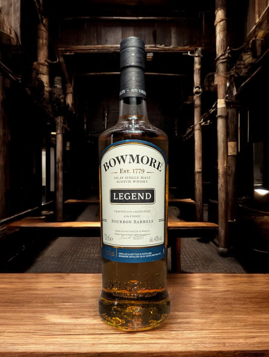Eine Flasche Bowmore Legend Whisky in einer klassischen Destilleriehalle, umgeben von Holzbalken, Steinwänden und Whiskyfässern. Die schlichte, elegante Flasche mit ihrem charakteristischen Etikett fügt sich harmonisch in die traditionelle, maritime Atmosphäre der Bowmore-Destillerie ein.