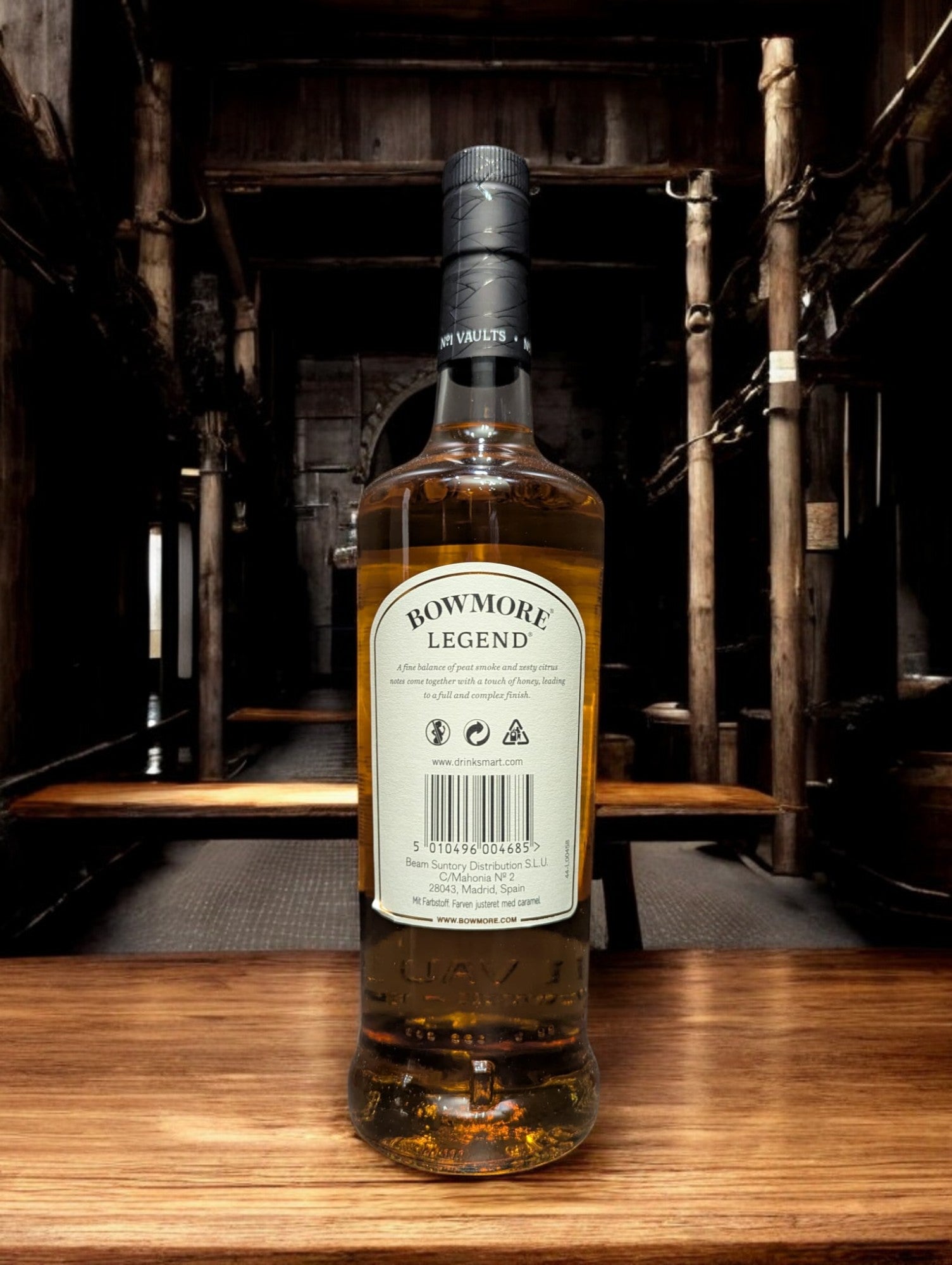 Eine Flasche Bowmore Legend Whisky in einer klassischen Destilleriehalle, umgeben von Holzbalken, Steinwänden und Whiskyfässern. Die schlichte, elegante Flasche mit ihrem charakteristischen Etikett fügt sich harmonisch in die traditionelle, maritime Atmosphäre der Bowmore-Destillerie ein.