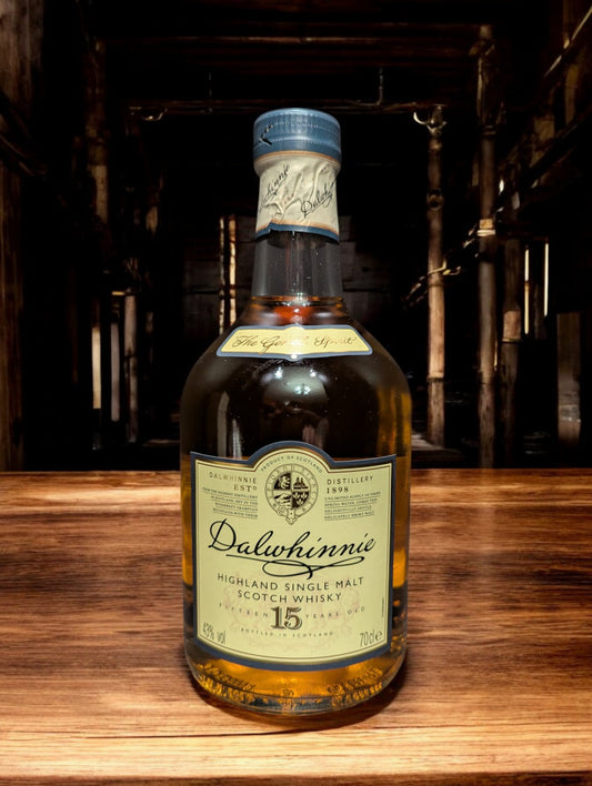 Eine Flasche Dalwhinnie 15 Jahre Single Malt Scotch Whisky steht aufrecht vor einem dunkelgrauen Hintergrund. Die elegante, klare Glasflasche zeigt ein cremefarbenes Etikett mit goldenen Akzenten und schwarzer Schrift. Der goldene Whisky schimmert im Licht und wird von einem markanten, kugelförmigen Flaschenhals mit einem schwarzen Verschluss gekrönt. Die gesamte Szene wirkt edel und zeitlos, was die hohe Qualität des Whiskys unterstreicht.