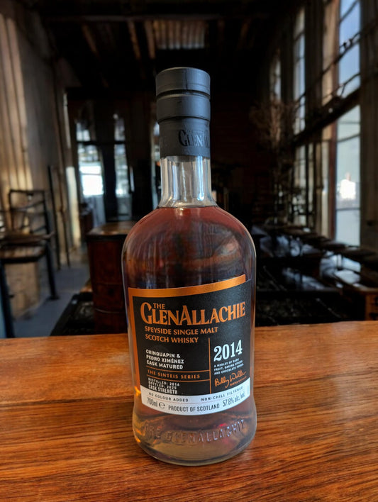 GlenAllachie Sinteis Part I Flasche auf einem niedrigen Holztisch, im Hintergrund eine dunkle Brennerei mit alten Holzfässern und kupfernen Brennblasen.