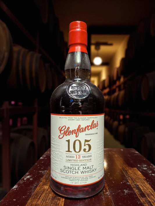 Glenfarclas 105 Hourglass Limited Edition Whisky bottle on weathered dark brown wooden table in traditional distillery setting with copper stills and oak barrels in shadowy background