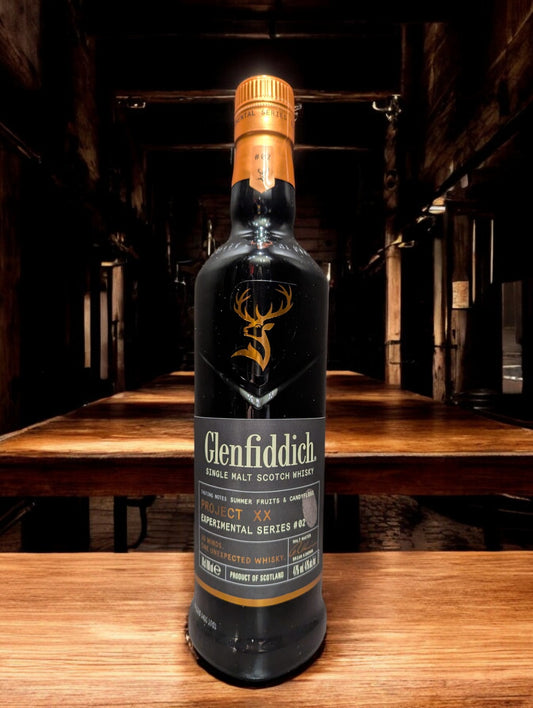 Ein elegantes Glenfiddich Project XX Whiskyfläschchen, präsentiert vor einem dunkelgrauen Hintergrund. Die Flasche besticht mit ihrem modernen Design: ein tiefschwarzes Etikett mit kupferfarbenem "XX"-Logo, das im Licht glänzt. Perfekt ausgeleuchtete Details betonen die hochwertige Verarbeitung, während der schlichte Hintergrund den Fokus auf das Produkt lenkt.