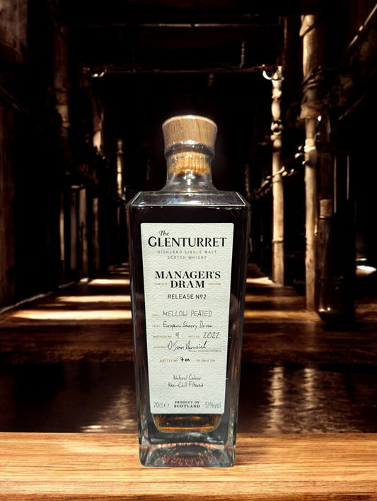 Eine Flasche Glenturret Manager's Dram No. 2 steht im Vordergrund, umrahmt von dunklem Holz und mit einem markanten, traditionellen Etikett. Der seltene schottische Single Malt Whisky verspricht mit seiner tiefen Bernsteinfarbe ein reiches Aromaprofil. Die limitierte Auflage wird durch eine individuelle Flaschennummer hervorgehoben, was Sammler und Kenner gleichermaßen anspricht. Der Hintergrund deutet auf eine exklusive Destillerie-Umgebung hin.