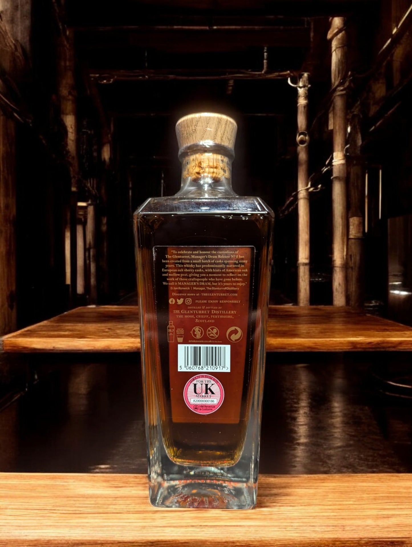 Eine Flasche Glenturret Manager's Dram No. 2 steht im Vordergrund, umrahmt von dunklem Holz und mit einem markanten, traditionellen Etikett. Der seltene schottische Single Malt Whisky verspricht mit seiner tiefen Bernsteinfarbe ein reiches Aromaprofil. Die limitierte Auflage wird durch eine individuelle Flaschennummer hervorgehoben, was Sammler und Kenner gleichermaßen anspricht. Der Hintergrund deutet auf eine exklusive Destillerie-Umgebung hin.