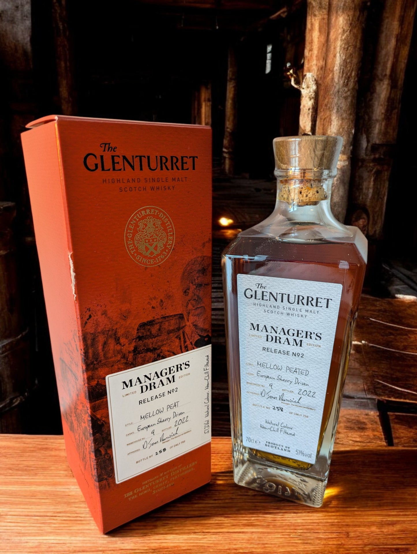 Eine Flasche Glenturret Manager's Dram No. 2 steht im Vordergrund, umrahmt von dunklem Holz und mit einem markanten, traditionellen Etikett. Der seltene schottische Single Malt Whisky verspricht mit seiner tiefen Bernsteinfarbe ein reiches Aromaprofil. Die limitierte Auflage wird durch eine individuelle Flaschennummer hervorgehoben, was Sammler und Kenner gleichermaßen anspricht. Der Hintergrund deutet auf eine exklusive Destillerie-Umgebung hin.