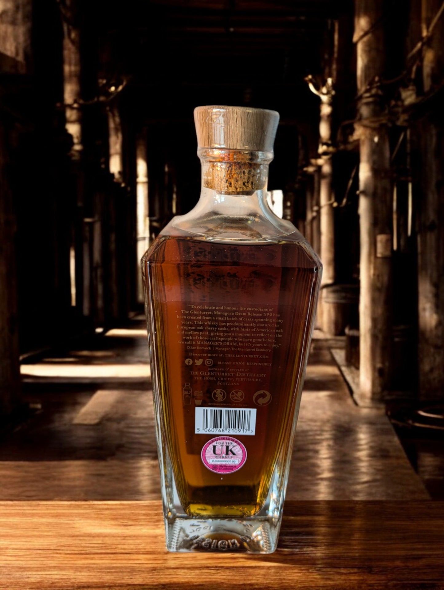 Eine Flasche Glenturret Manager's Dram No. 2 steht im Vordergrund, umrahmt von dunklem Holz und mit einem markanten, traditionellen Etikett. Der seltene schottische Single Malt Whisky verspricht mit seiner tiefen Bernsteinfarbe ein reiches Aromaprofil. Die limitierte Auflage wird durch eine individuelle Flaschennummer hervorgehoben, was Sammler und Kenner gleichermaßen anspricht. Der Hintergrund deutet auf eine exklusive Destillerie-Umgebung hin.