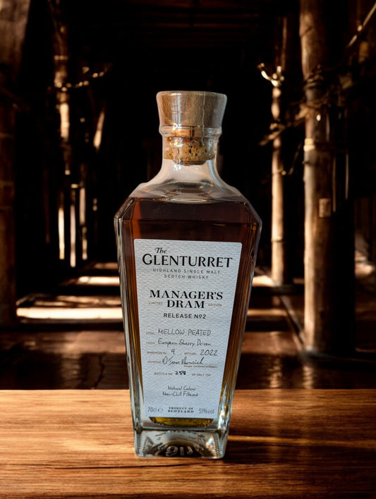 Eine Flasche Glenturret Manager's Dram No. 2 steht im Vordergrund, umrahmt von dunklem Holz und mit einem markanten, traditionellen Etikett. Der seltene schottische Single Malt Whisky verspricht mit seiner tiefen Bernsteinfarbe ein reiches Aromaprofil. Die limitierte Auflage wird durch eine individuelle Flaschennummer hervorgehoben, was Sammler und Kenner gleichermaßen anspricht. Der Hintergrund deutet auf eine exklusive Destillerie-Umgebung hin.