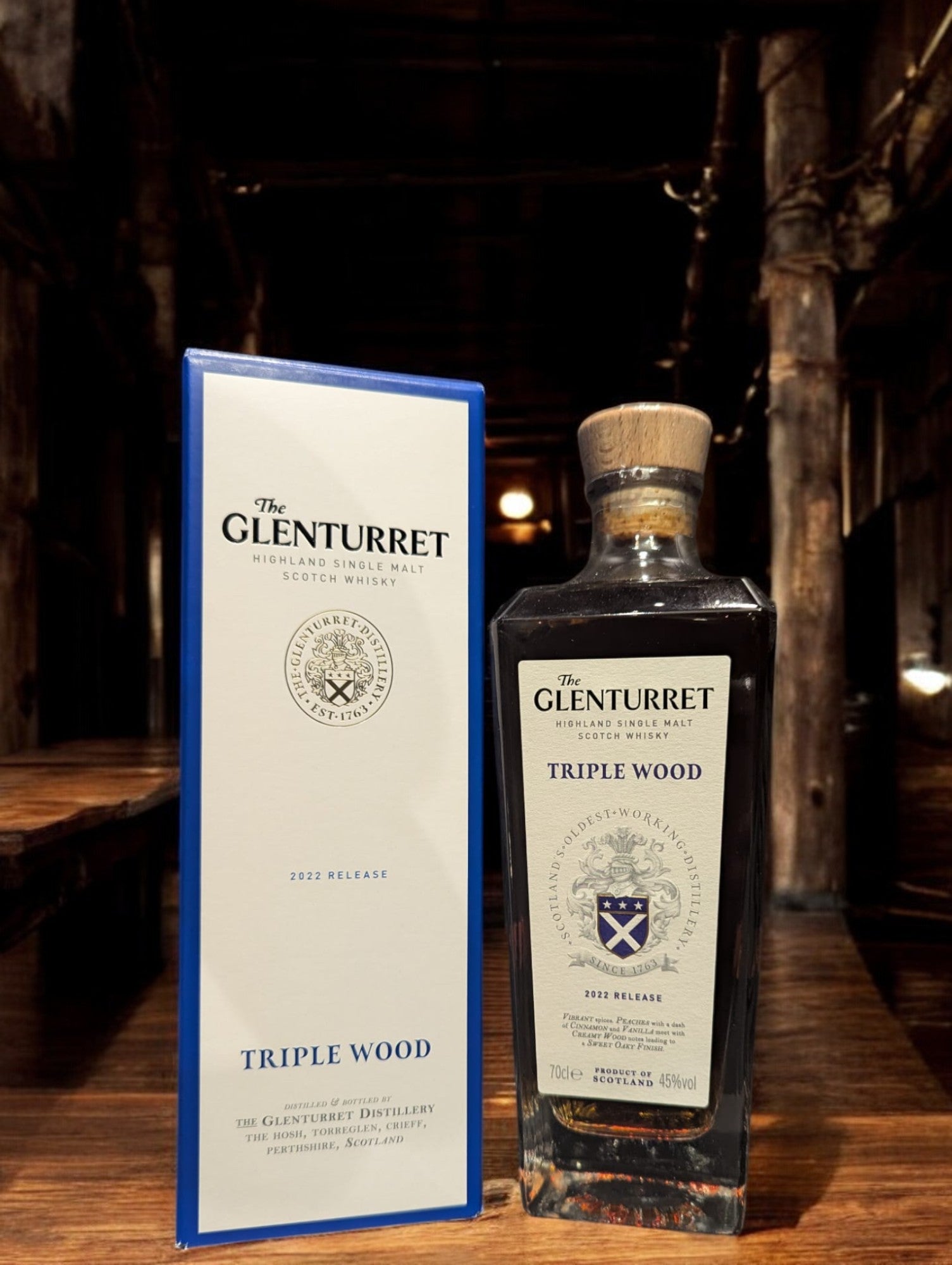 Glenturret Triple Wood Whiskyflasche elegant präsentiert mit sichtbaren Holzmaserungen im Hintergrund. Bernsteinfarbener Whisky reflektiert das Licht, während das Etikett die Reifung in drei verschiedenen Holzfässern hervorhebt. Dieses Bild betont die komplexe Handwerkskunst und die vielschichtigen Geschmacksnoten, die Liebhaber erwarten dürfen.