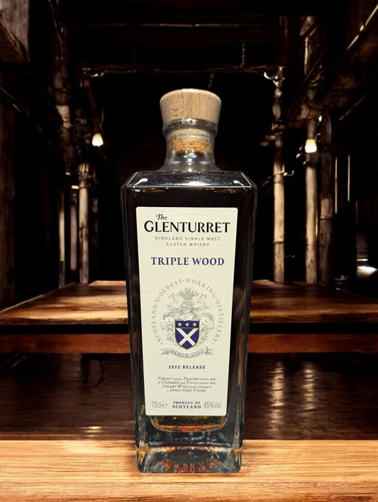 Glenturret Triple Wood Whiskyflasche elegant präsentiert mit sichtbaren Holzmaserungen im Hintergrund. Bernsteinfarbener Whisky reflektiert das Licht, während das Etikett die Reifung in drei verschiedenen Holzfässern hervorhebt. Dieses Bild betont die komplexe Handwerkskunst und die vielschichtigen Geschmacksnoten, die Liebhaber erwarten dürfen.