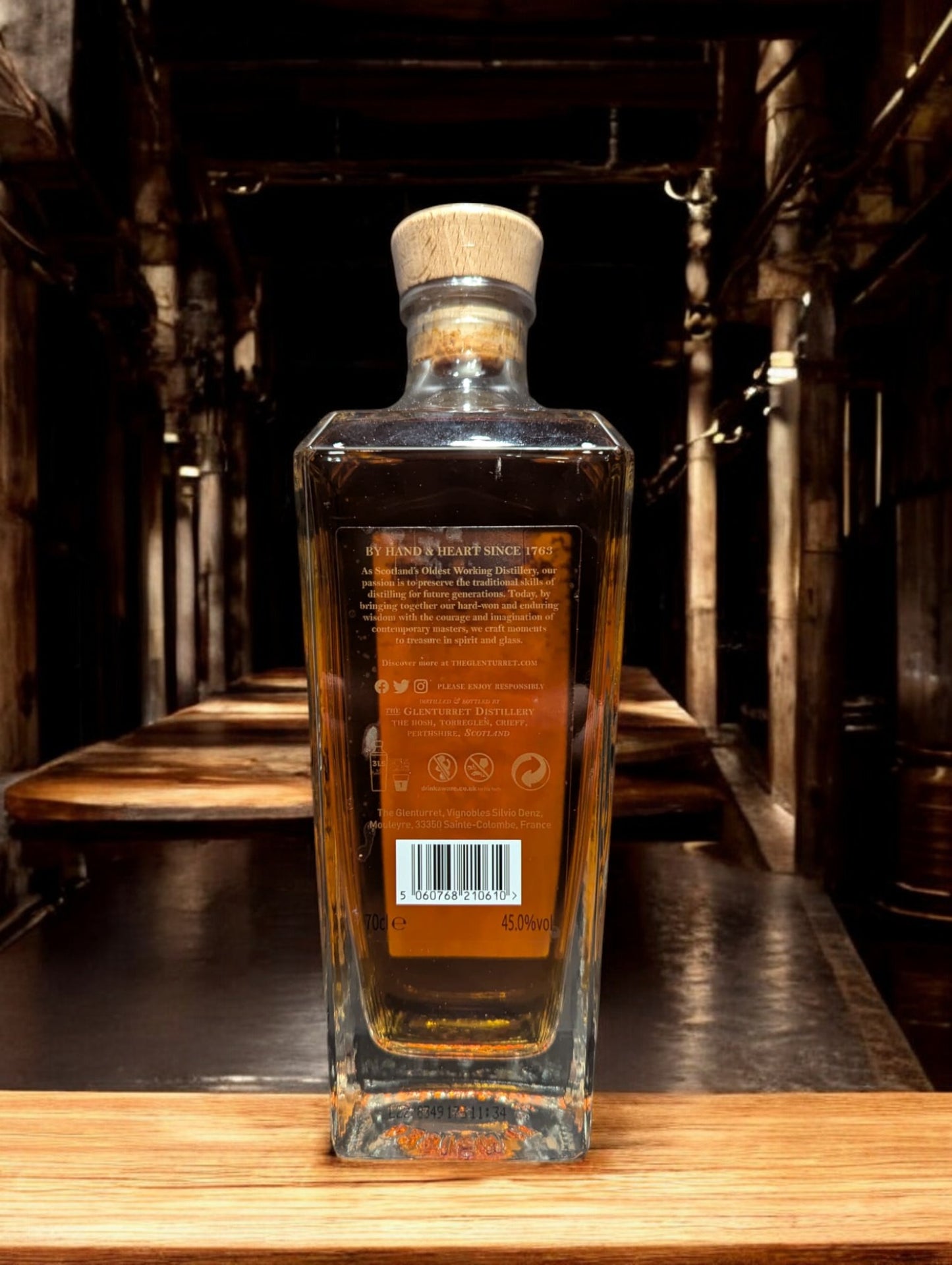 Glenturret Triple Wood Whiskyflasche elegant präsentiert mit sichtbaren Holzmaserungen im Hintergrund. Bernsteinfarbener Whisky reflektiert das Licht, während das Etikett die Reifung in drei verschiedenen Holzfässern hervorhebt. Dieses Bild betont die komplexe Handwerkskunst und die vielschichtigen Geschmacksnoten, die Liebhaber erwarten dürfen.