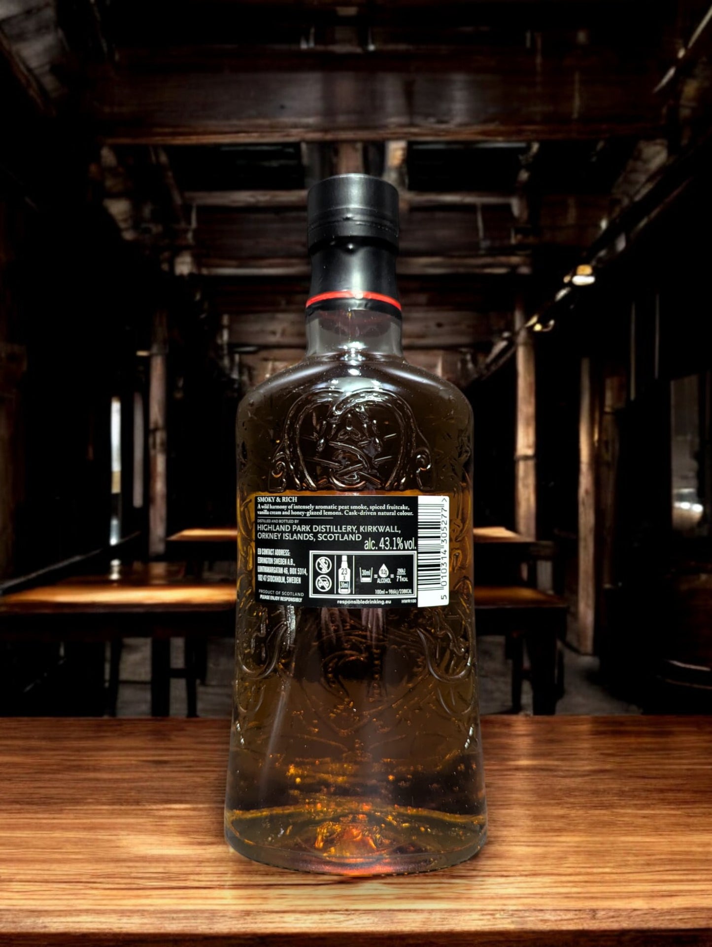 Eine Flasche Highland Park Dragon Legend Whisky in einer atmosphärischen Destilleriehalle, umgeben von rustikalem Holz und rauen Steinwänden. Die dunkle Flasche mit kunstvollem nordischen Drachen-Design sticht hervor und verleiht der Szenerie einen Hauch von mystischer Wikingertradition und handwerklicher Perfektion.