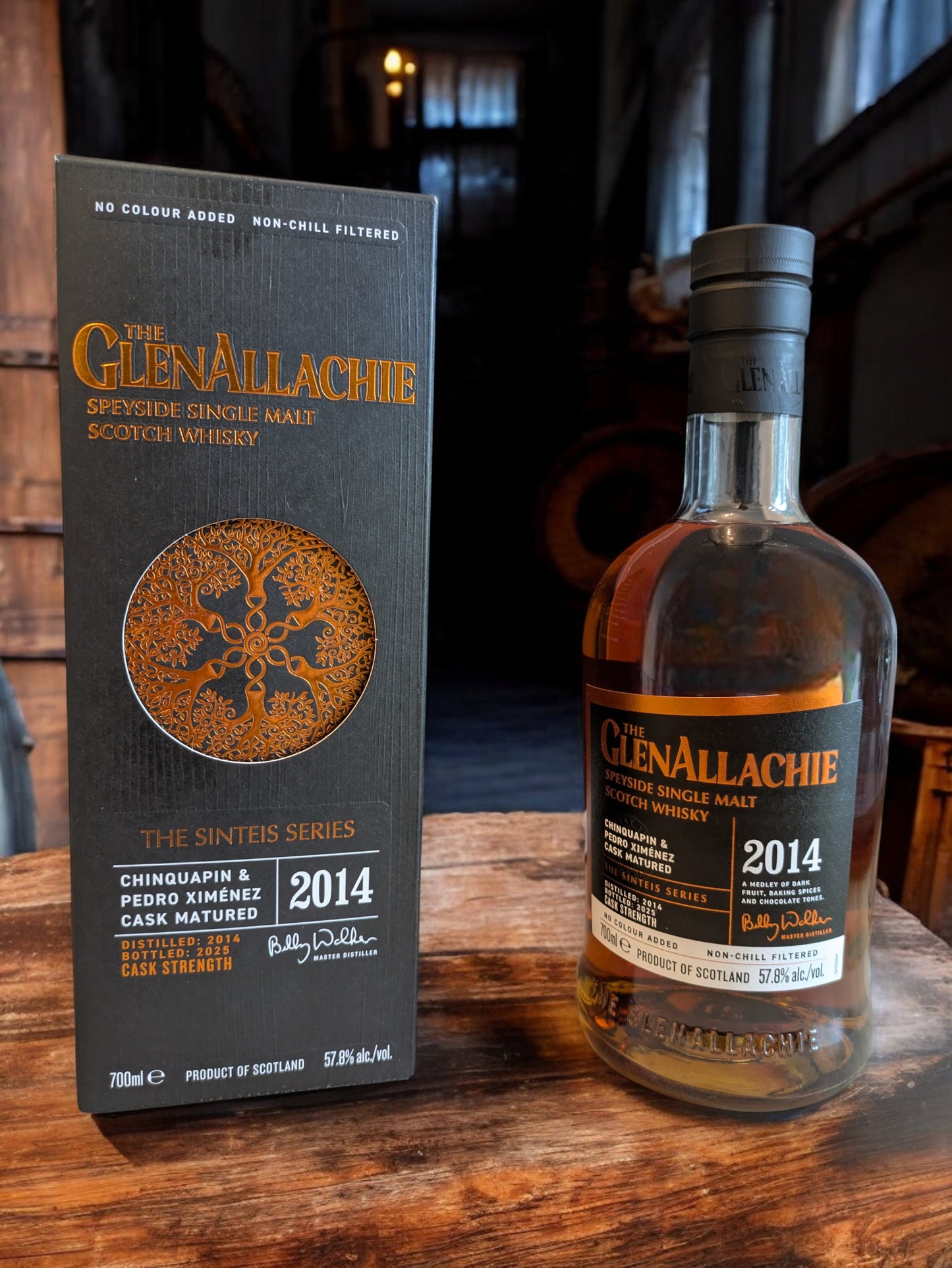 GlenAllachie Sinteis Part I Flasche auf einem niedrigen Holztisch, im Hintergrund eine dunkle Brennerei mit alten Holzfässern und kupfernen Brennblasen.