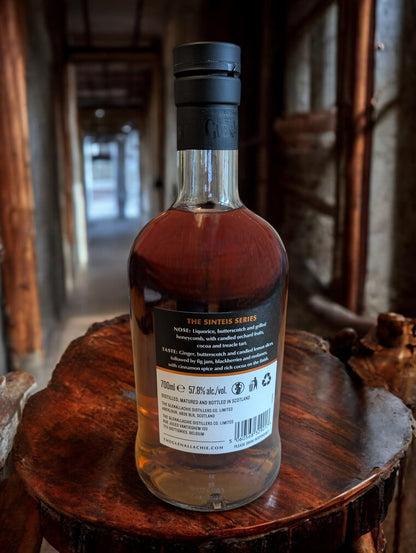GlenAllachie Sinteis Part I Flasche auf einem niedrigen Holztisch, im Hintergrund eine dunkle Brennerei mit alten Holzfässern und kupfernen Brennblasen.