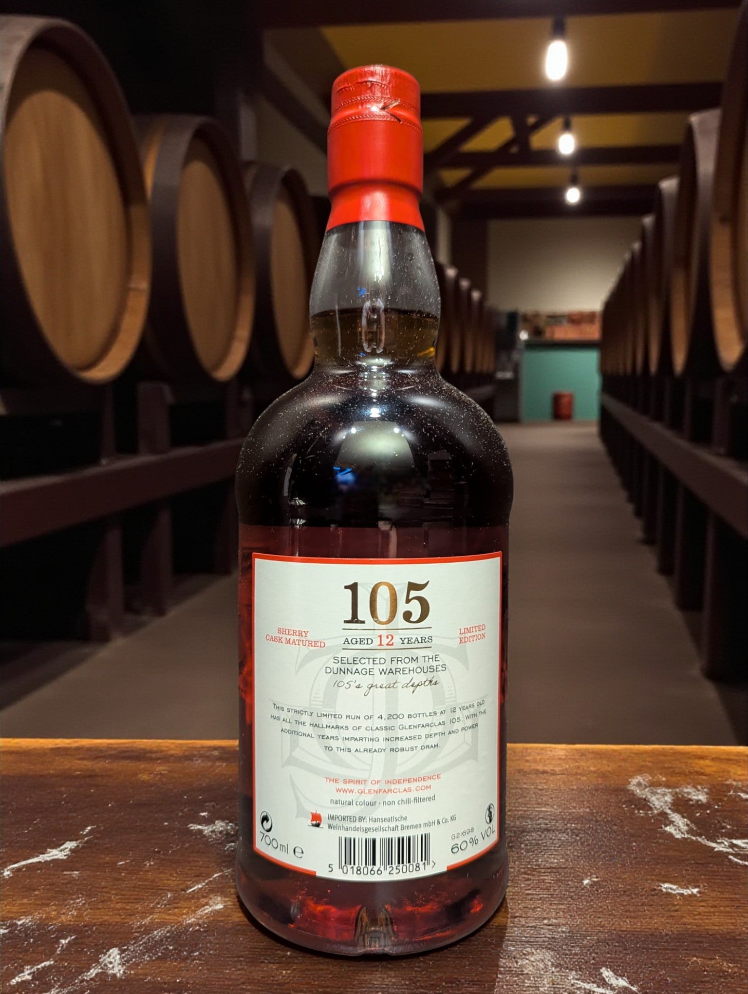 Glenfarclas 105 Hourglass Limited Edition Whisky bottle on weathered dark brown wooden table in traditional distillery setting with copper stills and oak barrels in shadowy background