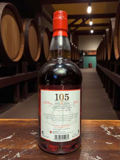 Glenfarclas 105 Hourglass Limited Edition Whisky bottle on weathered dark brown wooden table in traditional distillery setting with copper stills and oak barrels in shadowy background