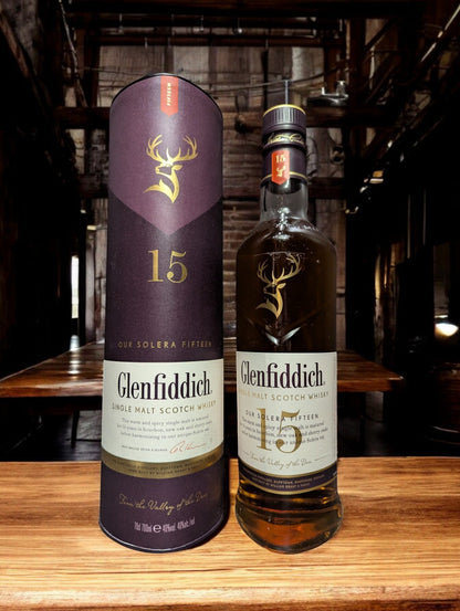 Ein Glenfiddich 15 Jahre Whisky in einer eleganten Flasche, umgeben von einer stilvollen Verpackung. Die goldene Farbe des Whiskys wird durch das Licht betont, während das Etikett die reiche Tradition und Qualität der Marke hervorhebt. Ideal präsentiert auf einem dunklen Holztisch mit einem passenden Whiskyglas, das einen Hauch von Luxus vermittelt.