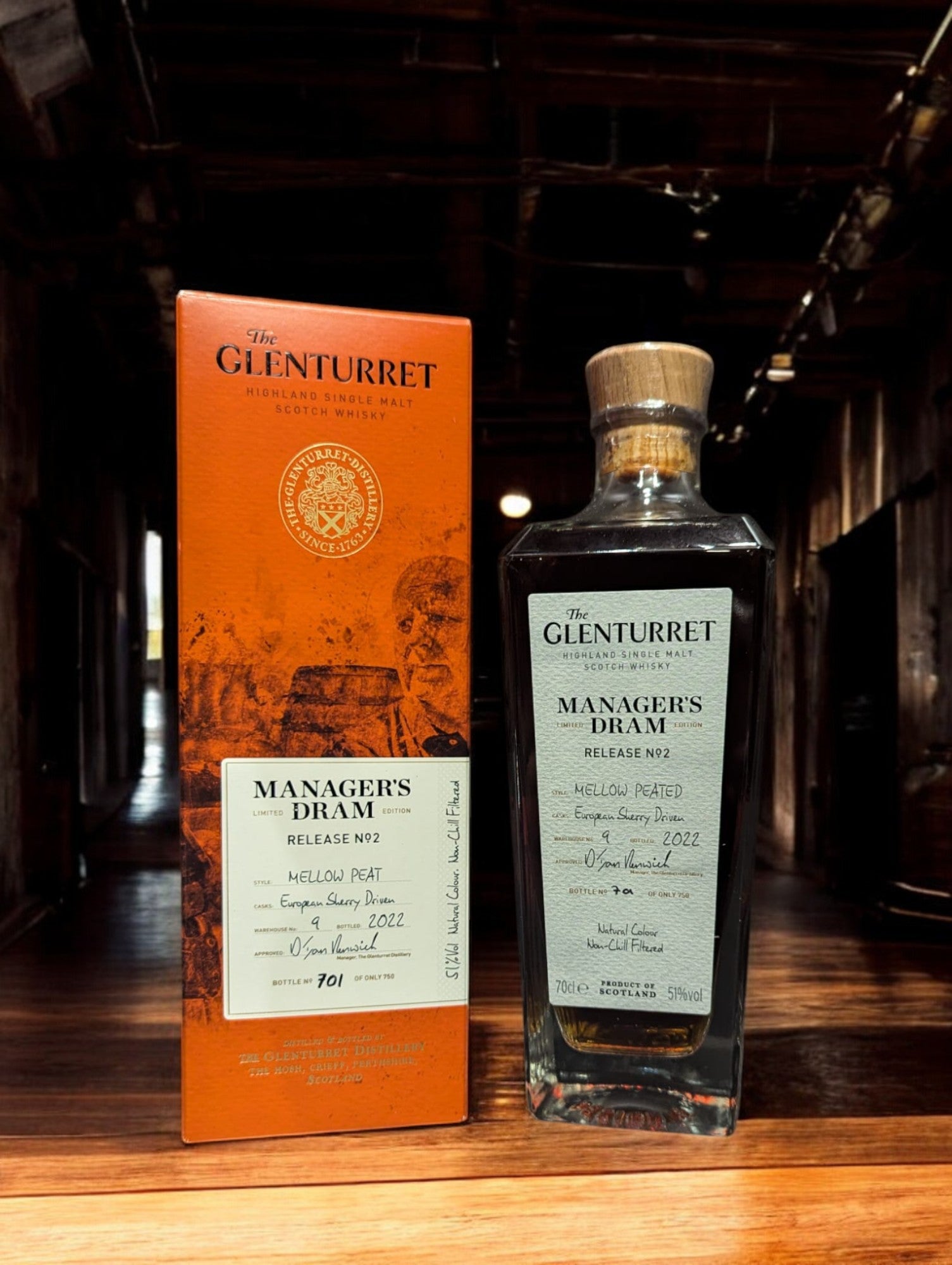 Eine Flasche Glenturret Manager's Dram No. 2 steht im Vordergrund, umrahmt von dunklem Holz und mit einem markanten, traditionellen Etikett. Der seltene schottische Single Malt Whisky verspricht mit seiner tiefen Bernsteinfarbe ein reiches Aromaprofil. Die limitierte Auflage wird durch eine individuelle Flaschennummer hervorgehoben, was Sammler und Kenner gleichermaßen anspricht. Der Hintergrund deutet auf eine exklusive Destillerie-Umgebung hin.
