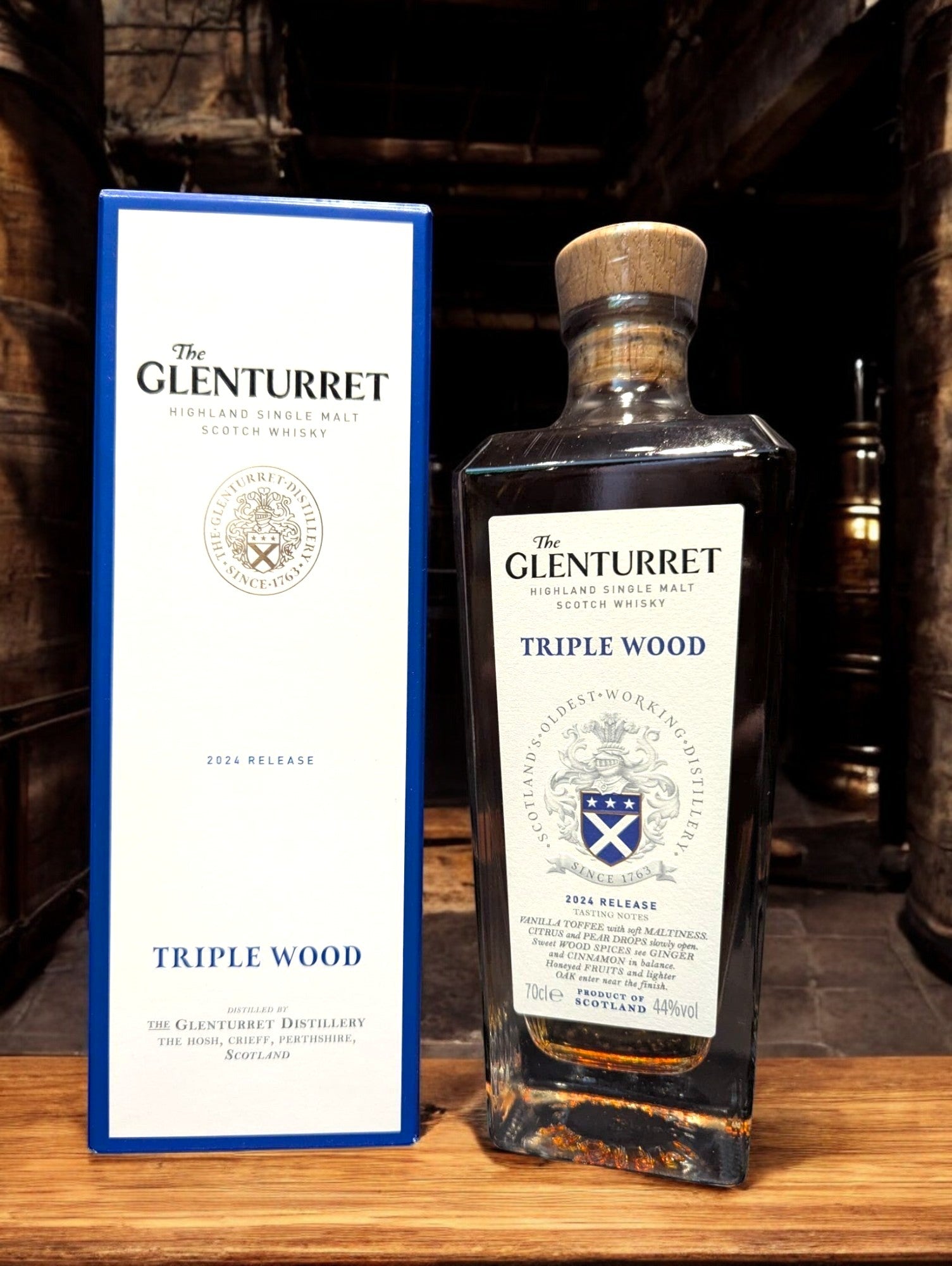 Glenturret Triple Wood Whiskyflasche elegant präsentiert mit sichtbaren Holzmaserungen im Hintergrund. Bernsteinfarbener Whisky reflektiert das Licht, während das Etikett die Reifung in drei verschiedenen Holzfässern hervorhebt. Dieses Bild betont die komplexe Handwerkskunst und die vielschichtigen Geschmacksnoten, die Liebhaber erwarten dürfen.