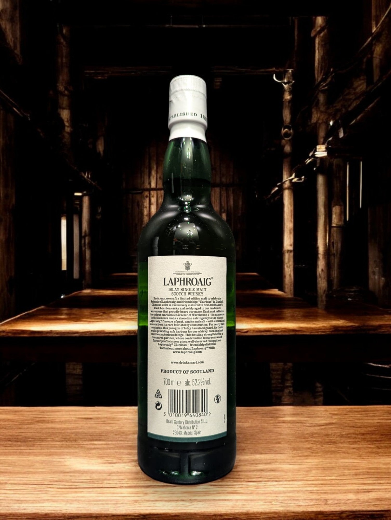 Flasche Laphroaig Cairdeas 2022 Warehouse 1 Edition Whisky mit markantem, dunklem Etikett und Siegel, präsentiert vor Fässern im Lagerhausstil, die die Herkunft und Exklusivität dieser limitierten Abfüllung betonen.