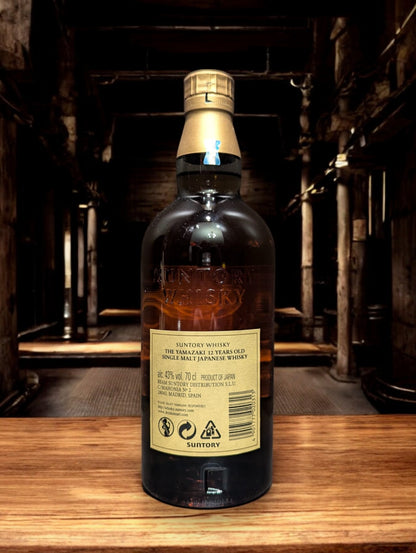 Detailaufnahme der Suntory Yamazaki Whiskyflasche, umgeben von Eichenfässern im Lagerhaus. Das warme Licht unterstreicht die goldbraune Farbe des edlen Single Malt Whiskys. Der Name Yamazaki prangt stolz auf dem Etikett, was auf die traditionsreiche Whiskybrennerei in Japan hinweist. Die Szene vermittelt eine Atmosphäre von Qualität und Handwerkskunst, die in der Herstellung dieses renommierten Getränks steckt.