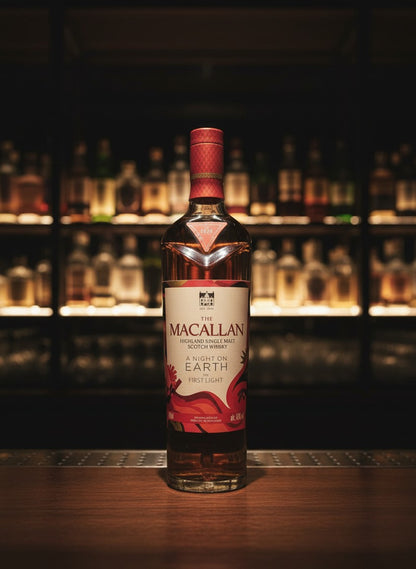 Whiskyquartier presents The Macallan A Night on Earth The First Light 2025 single malt Scotch whisky in a clear bottle with a red and white label, set in a distillery cellar.