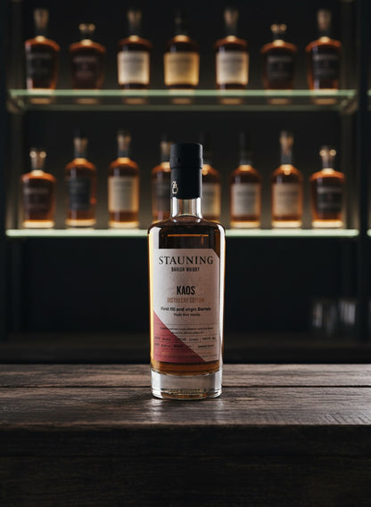 Whiskyquartier Stauning KAOS Triple Malt Whisky in 0.35L Flasche, bernsteinfarben, auf Sand mit Gräsern und blauem Himmel