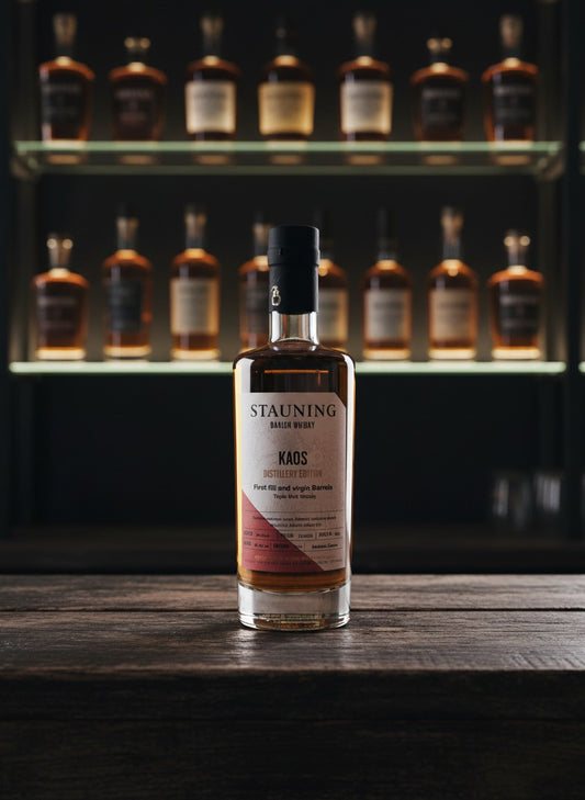 Whiskyquartier Stauning KAOS Triple Malt Whisky in 0.35L Flasche, bernsteinfarben, auf Sand mit Gräsern und blauem Himmel